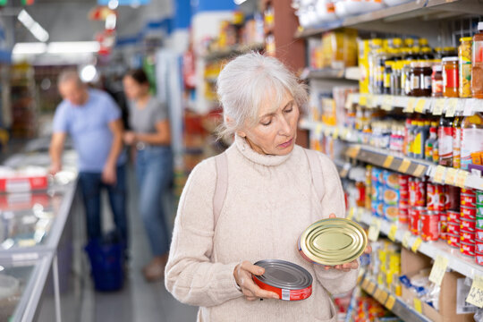 Focused interested elderly woman standing in grocery store aisle carefully reading nutritional information on tin cans while choosing right canned food.. - Powered by Adobe