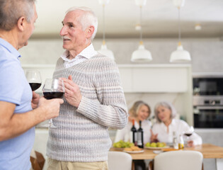 During house party, two elderly men stand at distance from dining area with glasses in their hands, discuss tips and tricks, share news, chatting