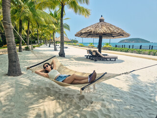 Brunette Russian girl relaxing in a beach hammock wearing denim mini skirt and t-shirt with sunglasses. Nha Trang beach with sunbeds and palm trees, Vietnam
