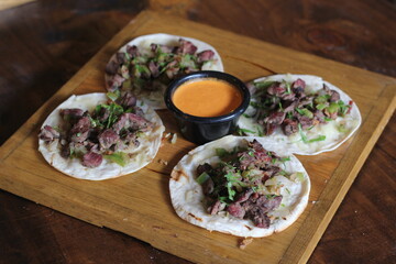 Four delightful tacos served on a wooden platter, showcasing tender meat with onions and cilantro, accompanied by a rich dipping sauce, perfect for street food lovers.