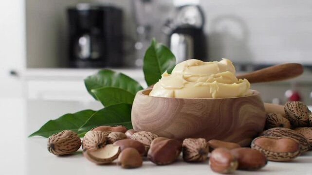 Shea Butter Cream in Wooden Bowl Surrounded by Nuts and Green Leaves in Clean Kitchen Setting