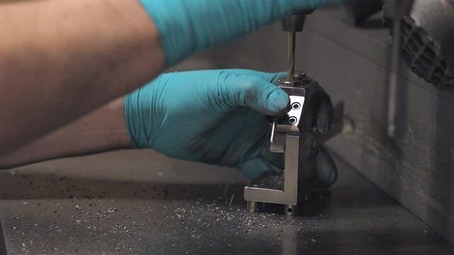 Close-up of a technician using a drill press and fixture to machine a small metal part, showcasing precision craftsmanship in a manufacturing setting.