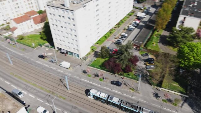Drone top-down shot of a tram arriving at a station in suburban Paris, showing houses, buildings, and a sunny parking lot - France