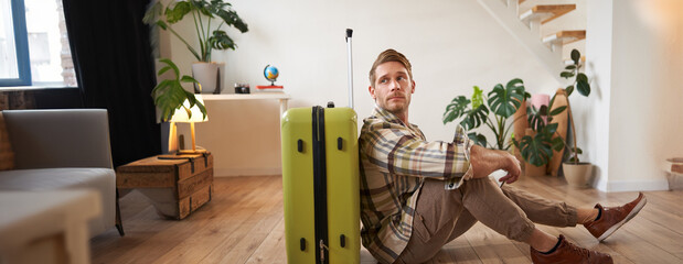 Portrait of male model with suitcase, sitting on floor, holding flight tickets and passport, going...