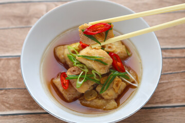 Close-up of steamed fish fillet held with chopsticks, garnished with sliced red chili and scallions. Captured above a white plate with broth, showcasing the tender texture and vibrant toppings.