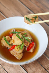 Portrait view of steamed fish fillet served in broth, with a piece lifted by chopsticks. Garnished with fried garlic, fresh scallions, and sliced chili. Captured on a white plate under natural light.