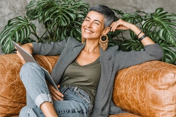 A stylish woman relaxes on a leather sofa, smiling with a tablet in hand and surrounded by lush green plants.
