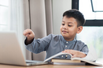 Asian young boy engaged in online learning, smiling while using laptop and studying with books. His focused expression reflects curiosity and enthusiasm for education