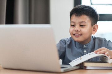 Asian young boy engaged in online learning, smiling while using laptop and reading book. setting is bright and modern, reflecting positive learning environment