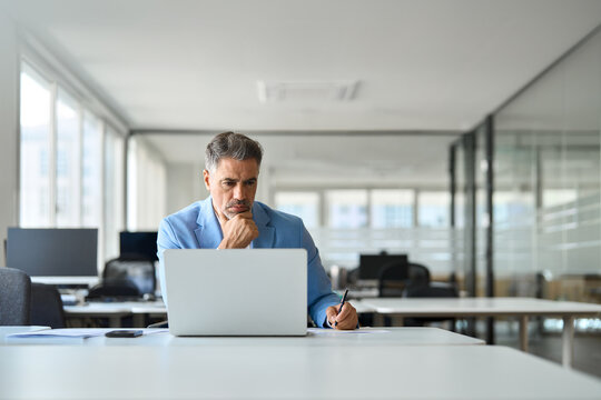 Busy professional executive manager looking at computer in office writing notes, serious old middle aged business man wearing suit working on laptop analyzing financial market, candid photo.
