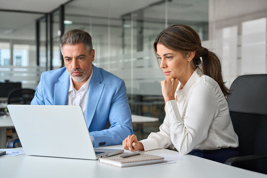Professional mature business man and woman leaders colleagues talking working on computer at workplace. Two busy professional executives company team working in corporate office using pc laptop.