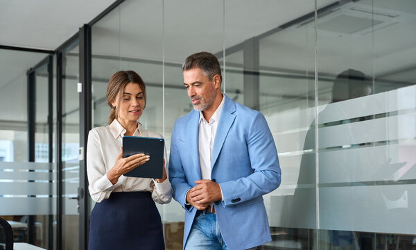 Executives team of two business people working talking using tablet tech walking in corporate office. Happy professional smiling businessman and businesswoman having conversation at work. Candid photo