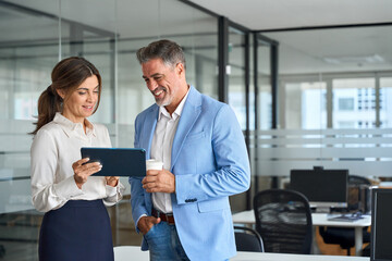 Executives team of two business people working together using tablet technology standing in corporate office. Happy professional smiling businessman and businesswoman having conversation at work.