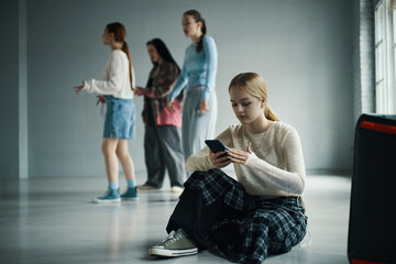 Fototapeta premium Teenager sitting on floor focused on smartphone with friends practicing dance in background in spacious room with natural light streaming through windows