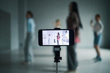 Group of dancers practicing routine while being filmed with smartphone on tripod. Focus on phone screen showing live recording of dance session, creating an engaging composition