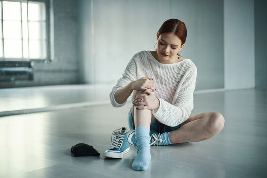Young woman sitting on floor stretching her leg in brightly lit dance studio. Reflective surface of studio floor and windows in background adding ambiance