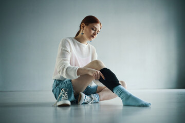 Caucasian woman preparing for sports activity while sitting on floor indoors, adjusting protective knee pad for comfort and safety. Wearing casual athletic attire and sneakers
