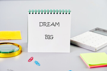 A notepad featuring the motivational phrase dream big sits on a clean workspace surrounded by colorful sticky notes, paper clips, and a magnifying glass, encouraging creativity and planning