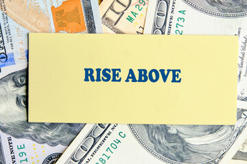 A card with the phrase rise above rests among various U.S. dollar bills. The composition highlights...