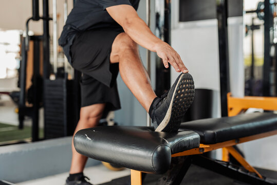 Closeup of anonymous man doing standing hamstring stretch with one foot raised on a gym bench, wearing athletic shoes.