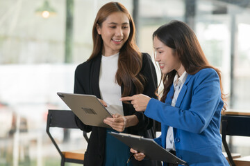 Two  Asian businesswomen holding iPads, holding graph papers, standing in front of the glass, talking and consulting with each other.