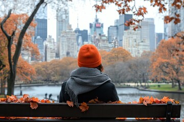 A person in an orange beanie sits on a park bench surrounded by autumn leaves. gazing at the New York City skyline with skyscrapers in the background