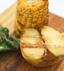 Grilled corn with potato and chili pepper on wooden cutting board.
