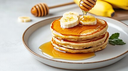 A stack of golden pancakes is drizzled with honey and topped with fresh banana slices on a plate, with bananas and a honey dipper in the background.