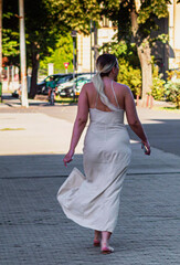 Attractive and fashionable young  woman wearing a long cream dress with thin straps walk in the city park. Sexy blonde girl in casual summer clothes on the city street alone. Rome, Italy, May 8, 2025
