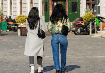 Naklejka premium Rear view of two stylish and elegant brunette young women walking on the street in the town square on a sunny spring day. Two attractive female friends walking in the downtown. Urban street life scene