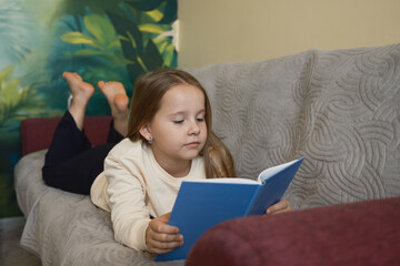 A young girl lies on a comfortable sofa, reading a blue book attentively. The relaxed and serene...