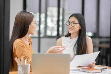 Two Asian businesswomen sit holding graph papers and discussing a new project.