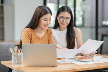Two Asian businesswomen holding graph documents, sitting and talking about work, looking happy...
