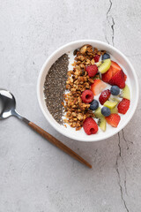 Top view of kefir bowl with granola, chia seeds, strawberry, raspberry, kiwi and blueberries, with wood and metal spoon on gray stone background.  Concept of healthy nutrition and freshness