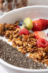 Detail of nutritious breakfast in white bowl: kefir, sliced fruit, golden granola and chia seeds. Blurred background with granola jar. Concept of mindful eating and healthy lifestyle