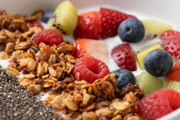 Close-up of fresh fruits, granola and chia seeds over kefir in a white bowl. Vibrant colors and crunchy texture. Concept of natural food, energy and vitality
