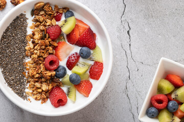 Top view of white bowl with kefir, granola, chia seeds and mixed fruits on grey stone surface. On the right, a small square bowl with more fruit. Concept of healthy breakfast and balanced eating