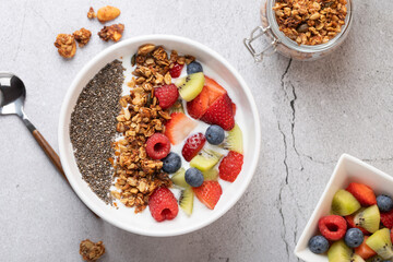 Top view of white bowl with kefir, granola, chia and fresh fruits. On the left, a metal spoon; on...