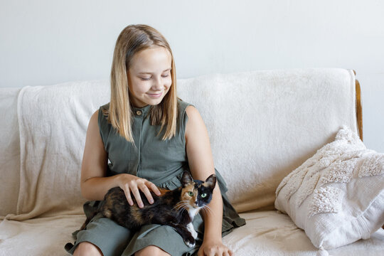 Portrait of young girl hugging her cat pet sitting on cosy sofa, relaxing at home. Child and domestic kitten indoor. - Powered by Adobe