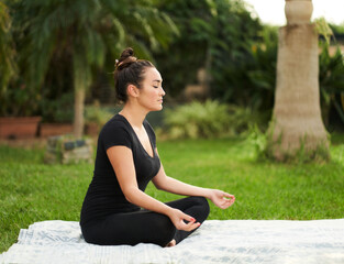 Fototapeta premium Young woman meditating in lotus position in a green garden