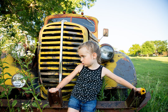 Young girl leans on antique yellow truck holding a flower