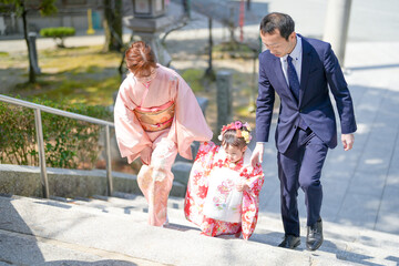 A 3-year-old Japanese girl in a red kimono walks up stairs with her parents-mom in a pink kimono,...