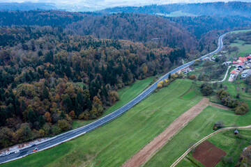New Road Construction Cutting Through Forest and Open Fields with Small Village and Hills in the Distance – Aerial View
