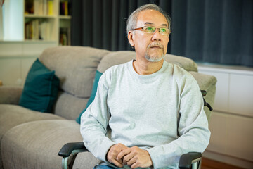 Fototapeta premium Asian senior man disabled sitting alone on wheelchair in living room looking out window at home and thinking, lonely elder sad old man retirement memory loss or nostalgia thought, feel depressed