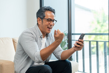 Fototapeta premium Handsome asian mature old man using smartphone on sofa in living room at home. Happy Portrait of cheerful smiling senior asian man holding cell phone. Mature People and lifestyle