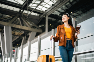 Happy young asian woman traveler using smartphone and holding boarding pass while standing at the...