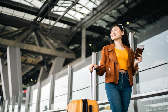 Happy young asian woman traveler using smartphone and holding boarding pass while standing at the airport terminal ready for vacation trip, smiling female tourist with confidently travel insurance
