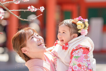 A 3-year-old Japanese girl in a red kimono celebrates Shichi-Go-San. Her mom, in a pink kimono, holds her as they smile at cherry blossoms. This shot shows a sweet moment between mother and daughter.