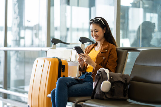 Young asian woman traveler using smartphone for checking flight information and playing social media app for shopping online while waiting with her luggage at the airport terminal..