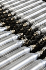 Neatly arranged silver gas cylinders with brass valves and engraved markings, displayed in an industrial setting. Cylinders show uniformity and technical precision